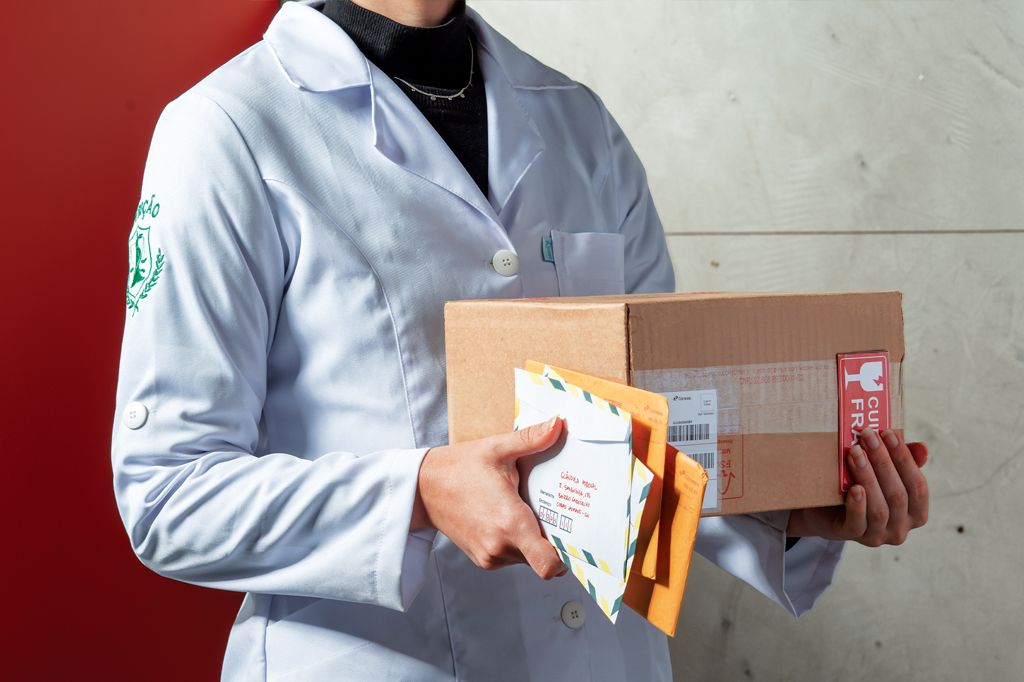 SI_477_Hormônios_4 Imagem de uma mulher, vestindo jaleco branco, carregando uma caixa de papelão e alguns envelopes.