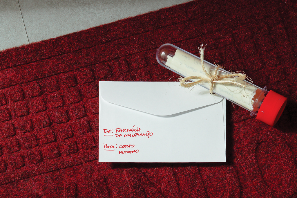 SI_477_Hormônios_5 Imagem de um envelope branco e um tubo de plástico em cima de um tapete vermelho.