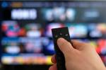 Imagem de uma mão segurando um controle remoto, em primeiro plano. Ao fundo, vê-se uma tela de TV desfocada.