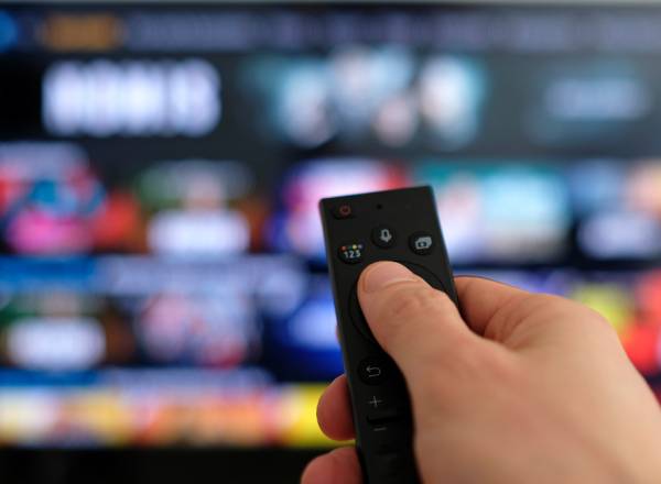 Imagem de uma mão segurando um controle remoto, em primeiro plano. Ao fundo, vê-se uma tela de TV desfocada.