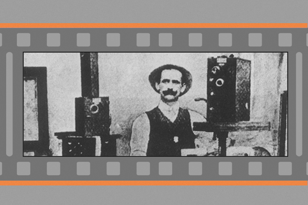 Fotografia, em fundo cinza, de Afonso Segreto (1898). V&ecirc;-se um homem de bigode e chap&eacute;u, cercado por antigas c&acirc;meras fotogr&aacute;ficas num est&uacute;dio.