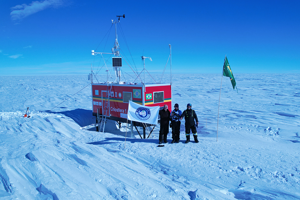 Fotografia da esta&ccedil;&atilde;o da miss&atilde;o criosfera ao lado de tr&ecirc;s cientistas segurando bandeira da miss&atilde;o.