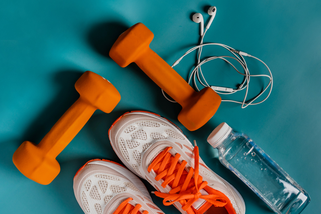 Imagem, vista de cima em fundo verde, de um par de tênis branco e laranja, uma garrafa d'água, um fone de ouvido e dois pesos pequenos para musculação.