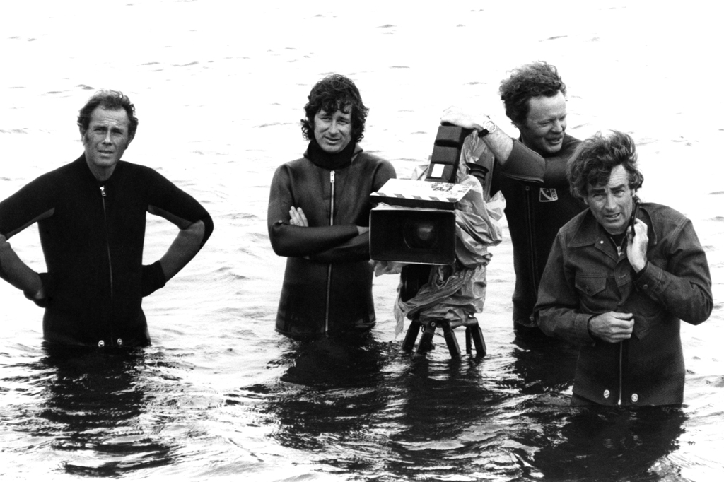 Imagem de quatro homens com &aacute;gua at&eacute; a cintura e equipamento de filmagem.