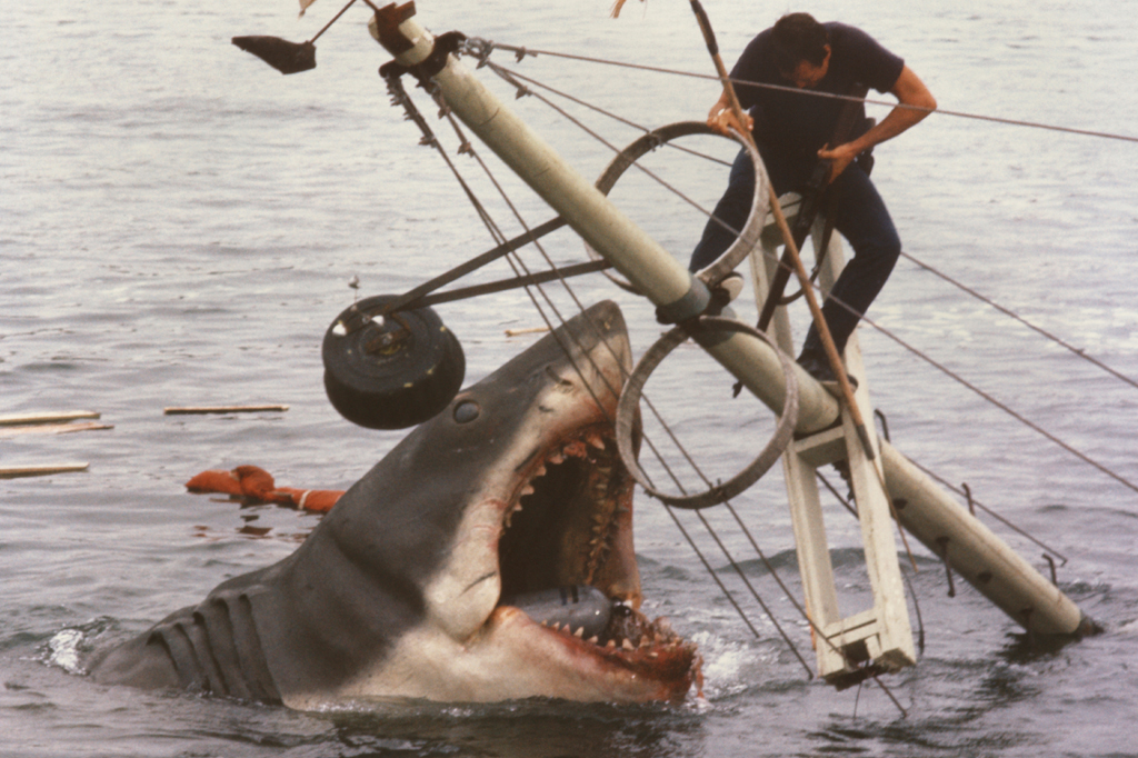 Imagem de um homem dependurado no mastro de barco afundando enfrentando um enorme tubar&atilde;o. Cena do filme 