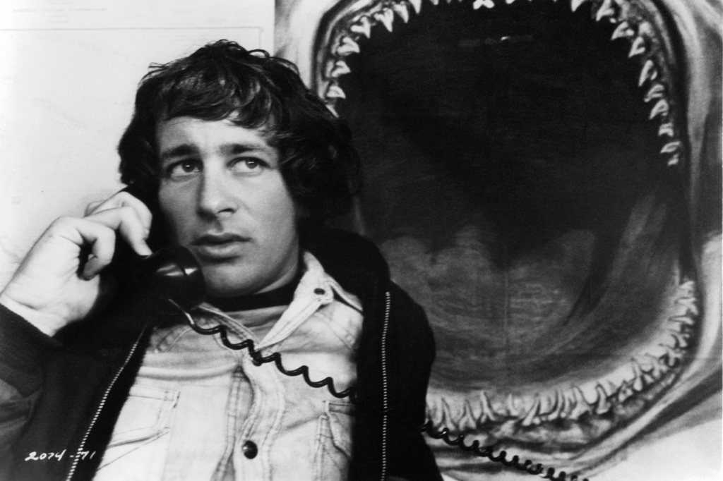 Imagem de um jovem falando ao telefone. Ao fundo, v&ecirc;-se um cartaz de um tubarn&atilde;o com a boca aberta.