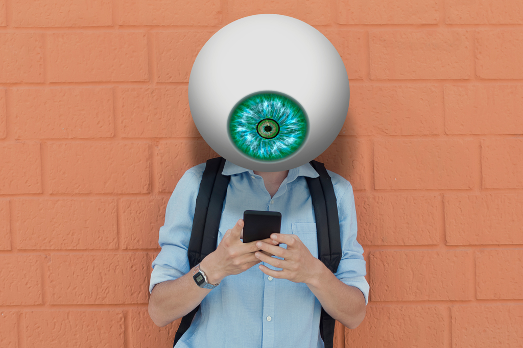 Imagem de um jovem com um gigantesco olho no lugar da cabeça checando o celular. Ao fundo, vê-se um muro de tijolos aparentes.