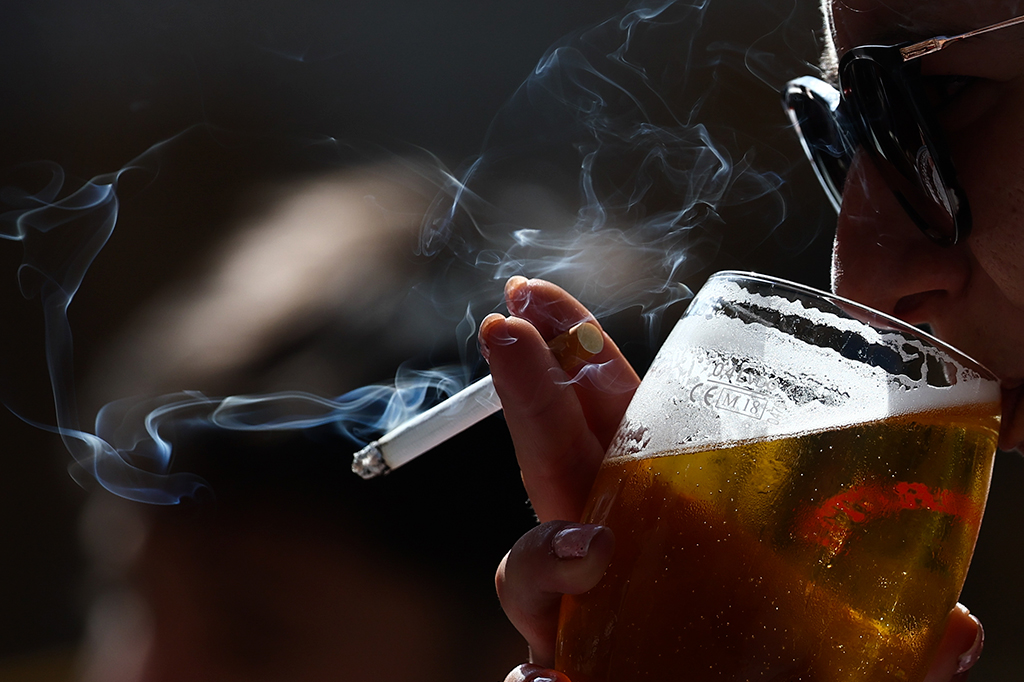 Fotografia de uma pessoa com um cigarro na mão bebe uma cerveja.