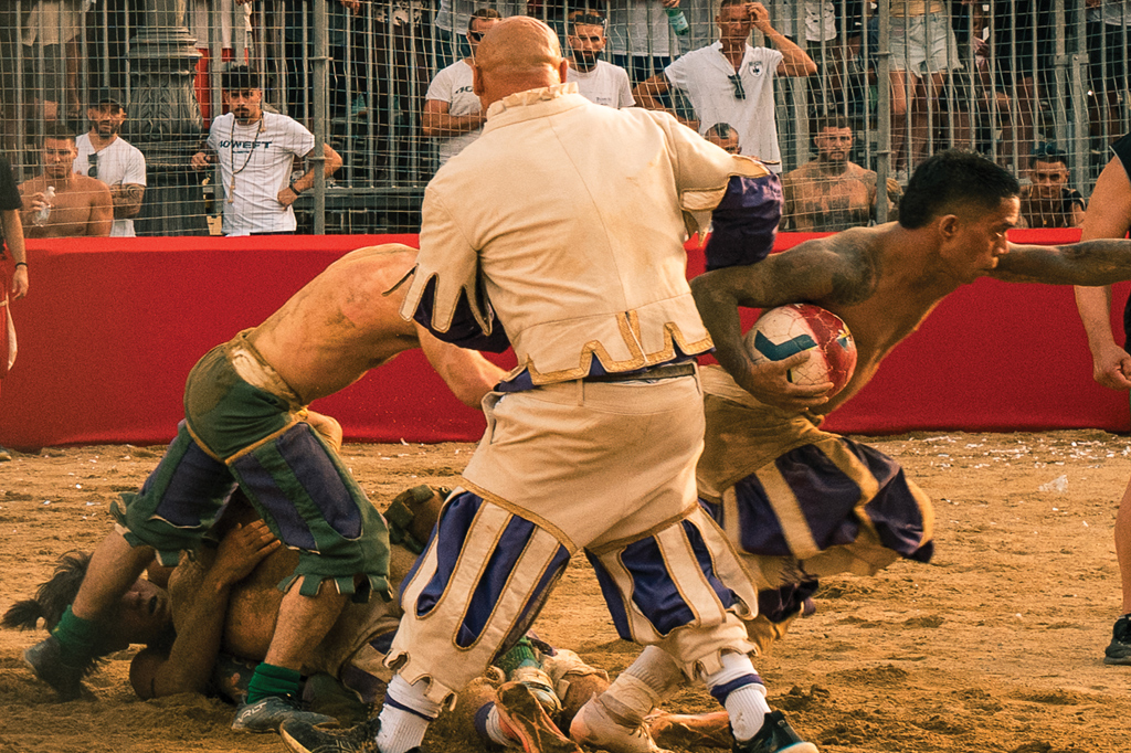 Fotografia de uma cena durante o torneio Calcio Storico Fiorentino de 2025: homens se agarram, sendo observados por um juiz.