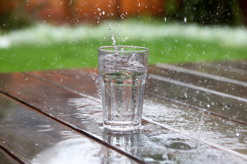 Imagem de um copo de vidro, sobre um piso de madeira, captando a água da chuva.
