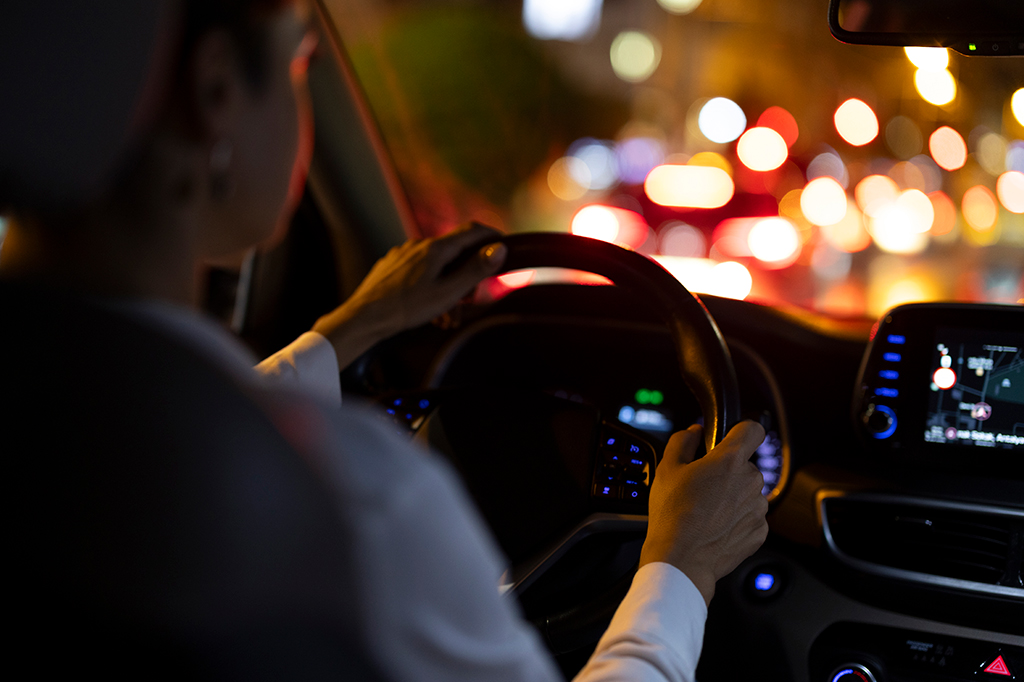 Fotografia de uma mulher dirigindo um carro à noite com luzes coloridas da cidade e tela de navegação iluminando o interior.
