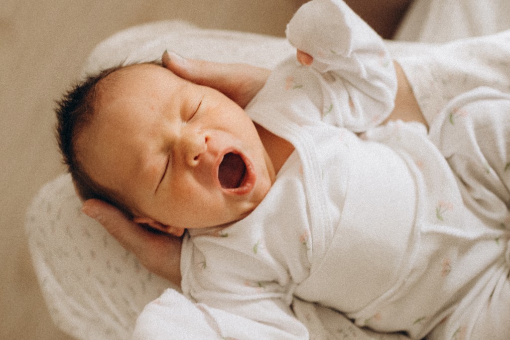 Bebê recém-nascido deitado de costas, bocejando com a boca aberta e os olhos fechados, vestido com um macacão branco estampado com pequenas flores cor-de-rosa, sendo segurado por mãos adultas em um fundo claro