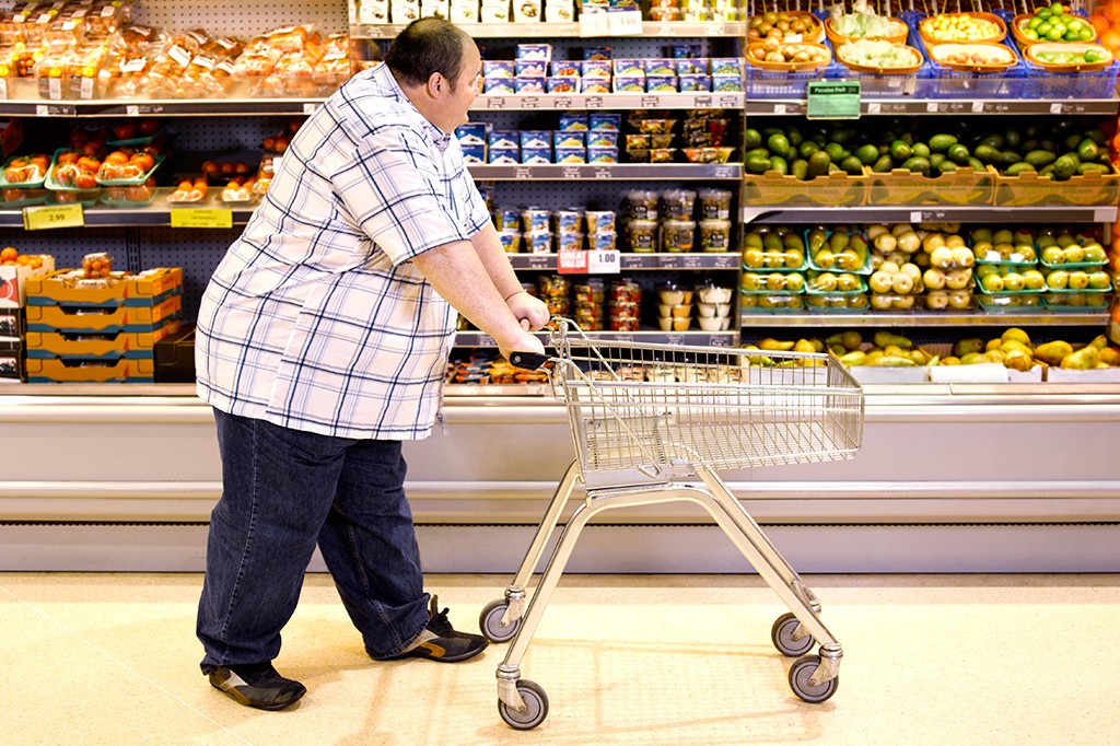 Homem de meia-idade, com sobrepeso, camisa xadrez e calça jeans, empurra um carrinho de compras vazio em um supermercado, passando por prateleiras de frutas, legumes e produtos enlatados
