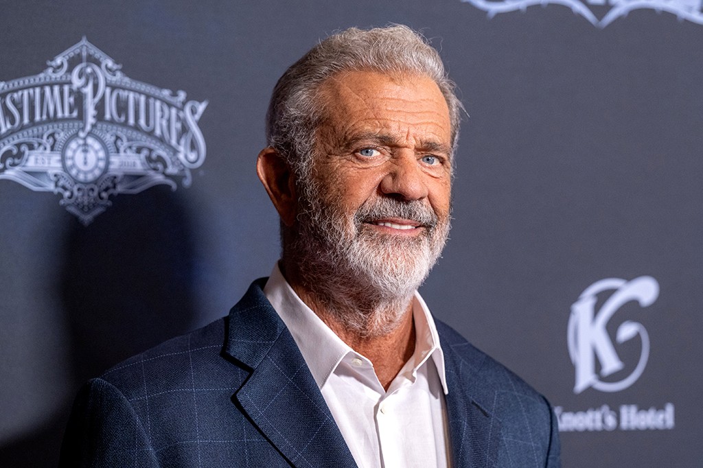 Mel Gibson, homem branco de cabelos grisalhos e barba, sorrindo levemente, vestindo blazer xadrez azul e camisa branca, em frente a um fundo escuro com logotipos