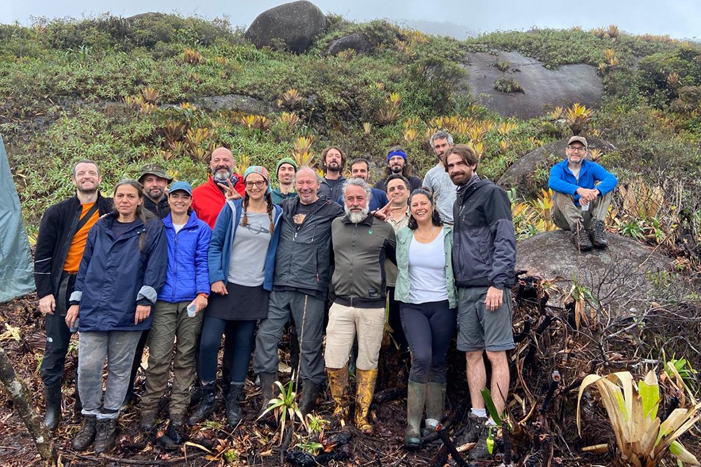 Grupo de 18 pessoas, incluindo homens e mulheres de diversas idades, sorrindo e posando para foto em trilha montanhosa. Vestem roupas e botas de trekking, alguns com capas de chuva. Um homem de azul est&aacute; sentado em uma rocha ao fundo. O cen&aacute;rio &eacute; de vegeta&ccedil;&atilde;o rasteira e rochas, com c&eacute;u nublado