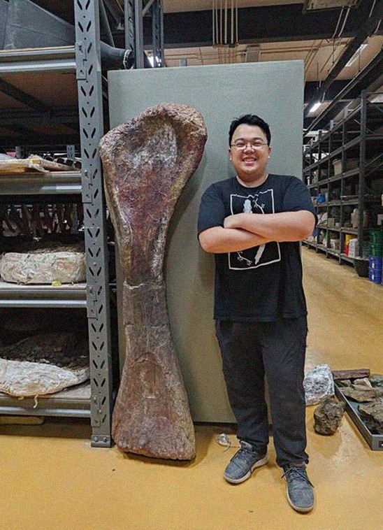Homem asi&aacute;tico sorridente, de &oacute;culos e camiseta preta, bra&ccedil;os cruzados, posa ao lado de um enorme osso f&oacute;ssil marrom, em um laborat&oacute;rio com prateleiras cheias de amostras