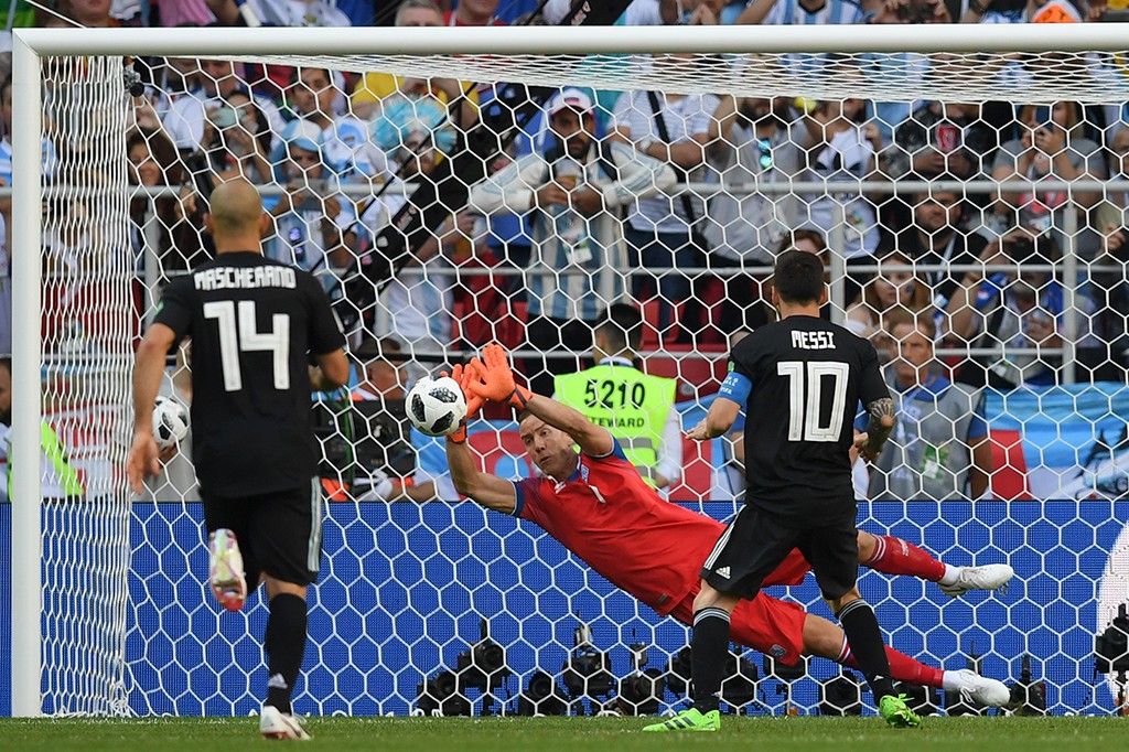 Goleiro em uniforme vermelho e luvas laranja salta para defender a bola, com Messi e Mascherano em camisas pretas observando a jogada, em um campo de futebol com torcida ao fundo