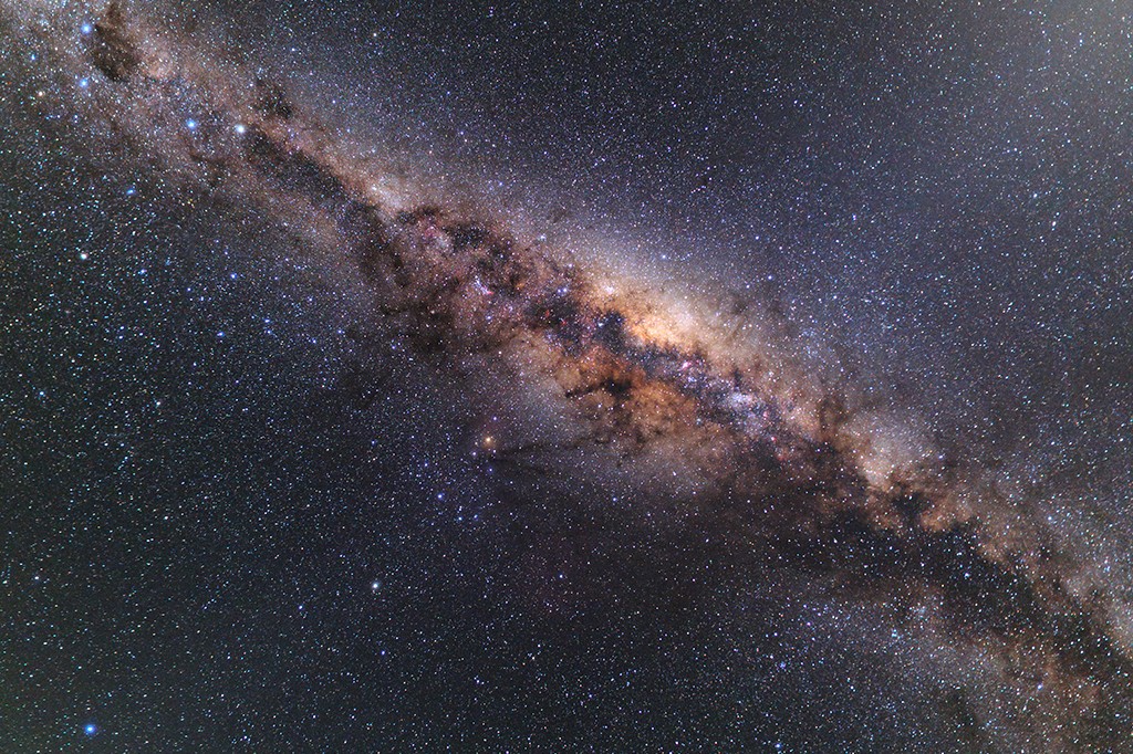 Via Láctea com bilhões de estrelas brilhantes, nuvens de gás e poeira em tons de marrom, laranja e roxo, atravessando um céu noturno escuro