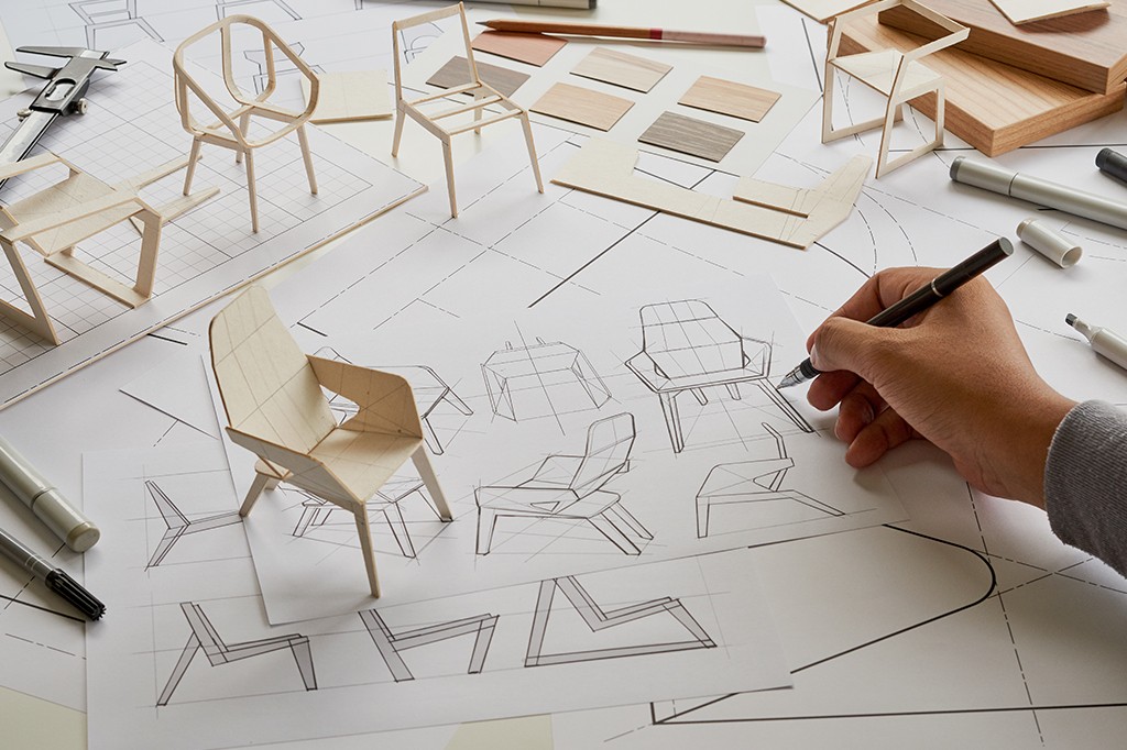 Mão desenhando esboços de cadeiras em papel quadriculado, com modelos em miniatura de madeira e amostras de materiais ao redor