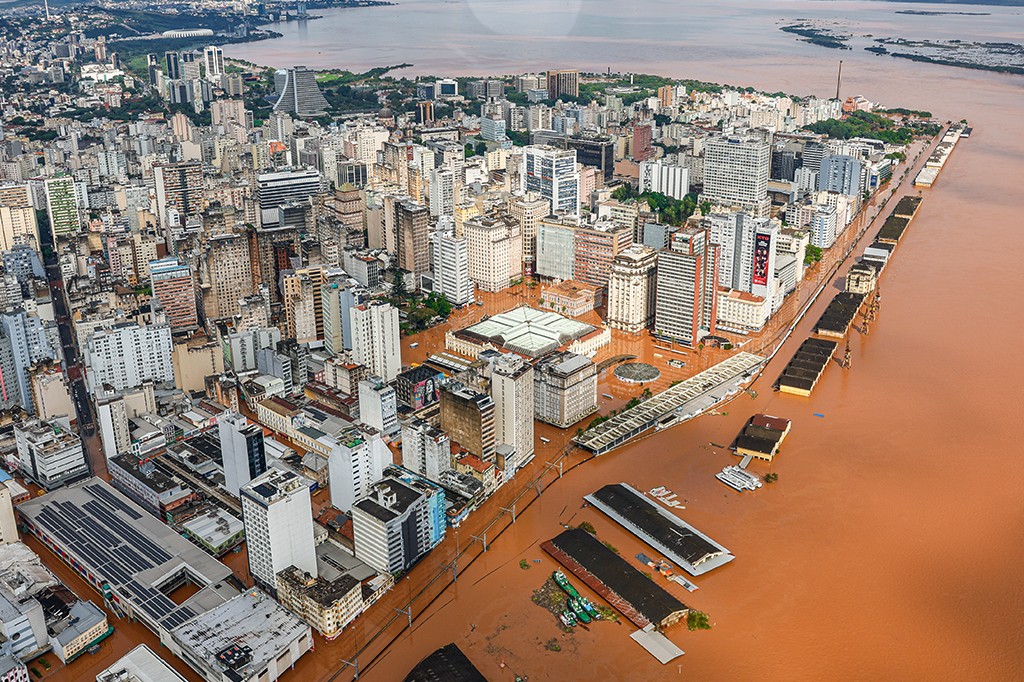 Vista aérea de Porto Alegre inundada, com prédios e ruas submersos em água barrenta. O centro da cidade e o cais do porto estão cobertos pela enchente, com apenas os telhados de algumas estruturas visíveis. O rio Guaíba, também marrom, se estende ao fundo, sob um céu nublado