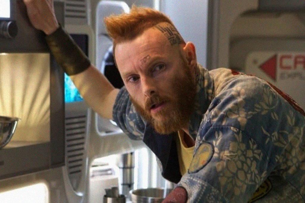 Homem ruivo com barba, moicano e tatuagens na cabeça, vestindo camisa azul floral e camiseta amarela, olhando para frente com expressão séria, apoiado em uma superfície metálica em um ambiente futurista