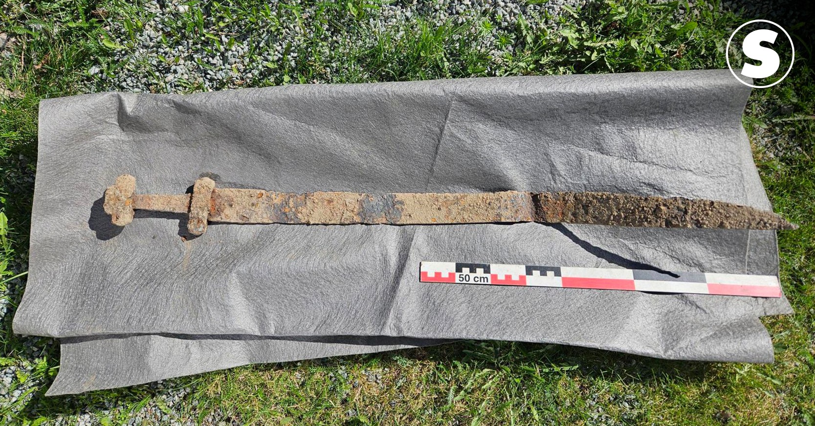 Espada antiga e enferrujada, com cabo em forma de T, sobre um tecido cinza escuro, com uma régua de 50 cm para escala, em um gramado com pedras