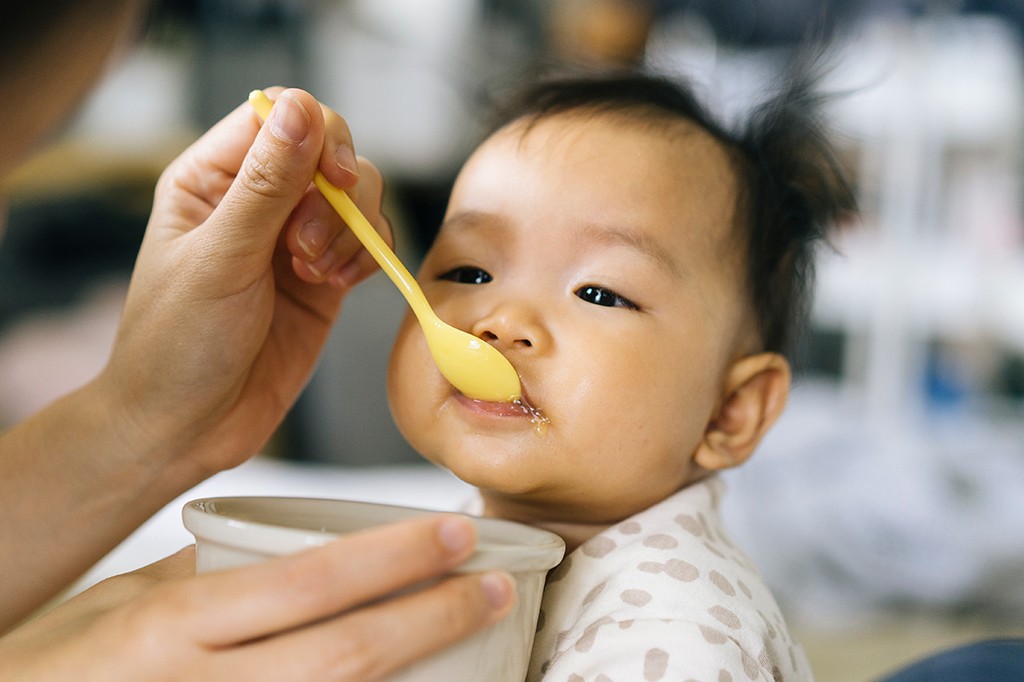 Mão adulta alimentando um bebê asiático com uma colher amarela, o bebê usa um macacão claro com bolinhas e tem um pouco de comida na boca