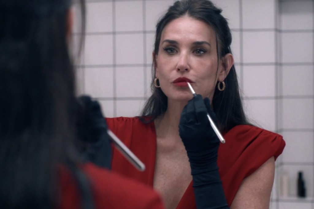 Mulher de cabelos escuros, vestindo um elegante vestido vermelho e luvas pretas, aplica batom vermelho com um pincel, olhando para seu reflexo no espelho. Ela usa brincos dourados e tem uma expressão concentrada. O fundo é uma parede de azulejos brancos com linhas cinzas