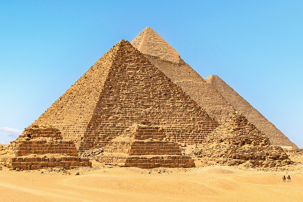 Três pirâmides de Gizé, no Egito, sob um céu azul claro, com a maior em primeiro plano e duas menores ao fundo. A areia dourada do deserto se estende à frente, e dois camelos com cavaleiros são visíveis à direita