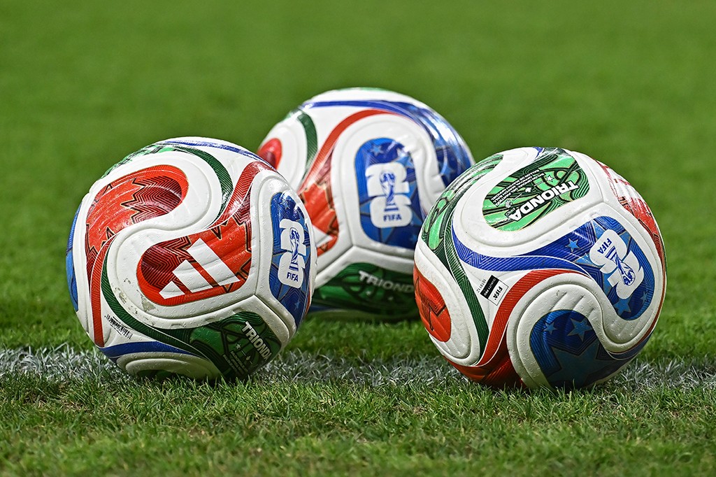 Três bolas de futebol Adidas Brazuca coloridas, com detalhes em vermelho, azul, verde e branco, sobre a grama verde de um campo, próximas a uma linha branca