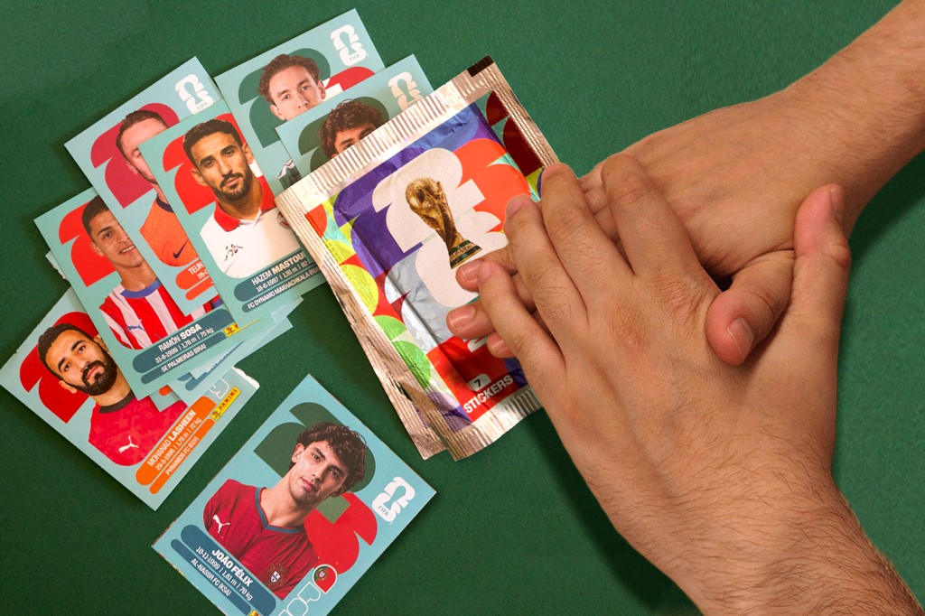 Mãos de uma pessoa abrindo um pacote de figurinhas da Copa do Mundo FIFA 2022, com várias figurinhas de jogadores espalhadas sobre uma superfície verde