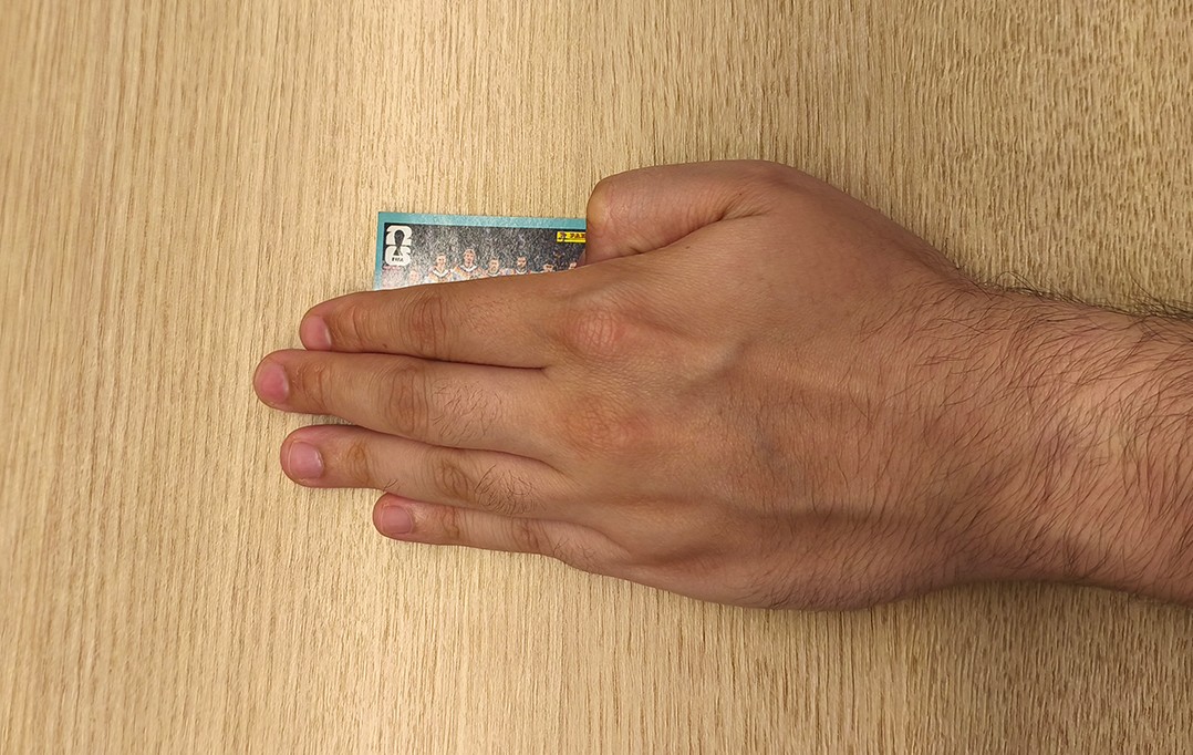 M&atilde;o masculina clara, com pelos no pulso, segurando um cromo de figurinhas da Panini com a imagem de um time de futebol, sobre uma superf&iacute;cie de madeira clara