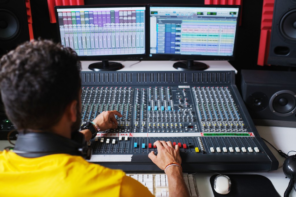 Imagem de um rapaz controlando uma mesa de som numa emissora de rádio.