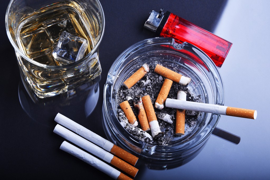 Image, vista de cima, de um cinzeiro com várias bitucas de cigarros e um copo de whisky com pedras de gelo. Ao lado vê-se um isqueiro vermelho e três cigarros intocados.