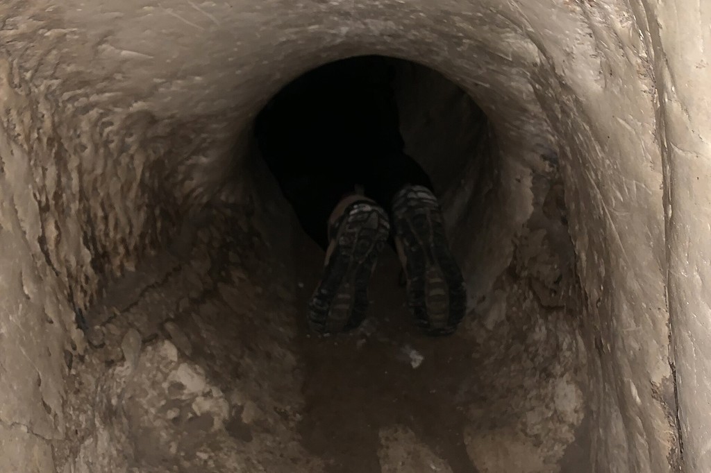 Pernas de uma pessoa com cal&ccedil;as escuras e t&ecirc;nis com solado sujo, entrando em um t&uacute;nel estreito e escuro de paredes rochosas irregulares.