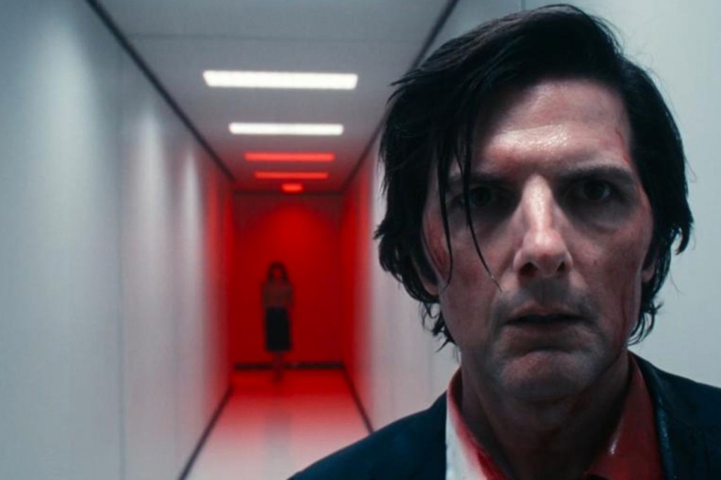 Homem de cabelo escuro e rosto sujo de sangue, com express&atilde;o de choque, olhando para frente. Ao fundo, um corredor iluminado em vermelho com uma figura feminina amea&ccedil;adora distante.