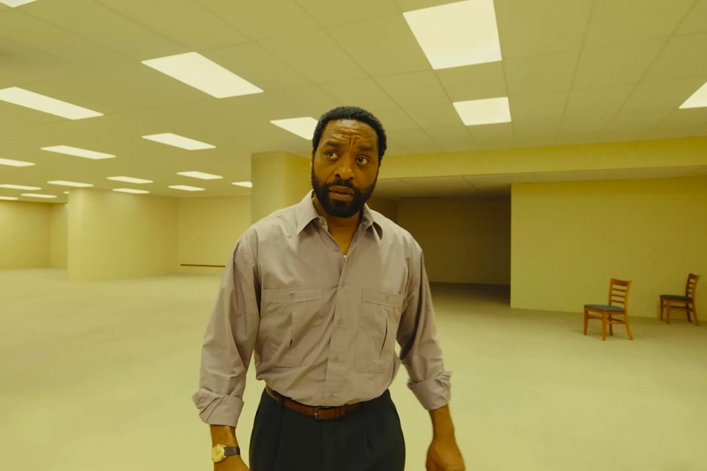Homem negro de barba, camisa cinza e cal&ccedil;a escura, com express&atilde;o confusa, em um ambiente vazio de paredes e ch&atilde;o amarelos, com luzes retangulares no teto e duas cadeiras de madeira ao fundo.