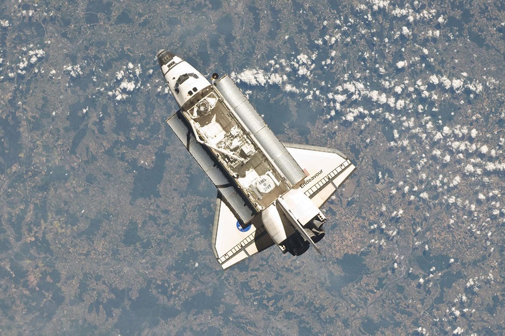 Ônibus espacial Endeavour, branco e cinza, visto de cima com a porta de carga aberta, orbitando a Terra com nuvens esparsas e paisagem terrestre visíveis abaixo
