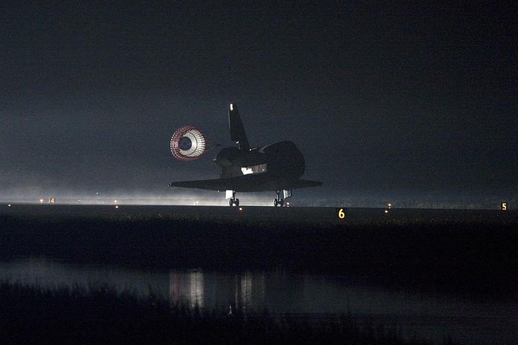 &Ocirc;nibus espacial escuro pousando em pista iluminada &agrave; noite, com paraquedas de freio aberto e reflexo na &aacute;gua &agrave; frente