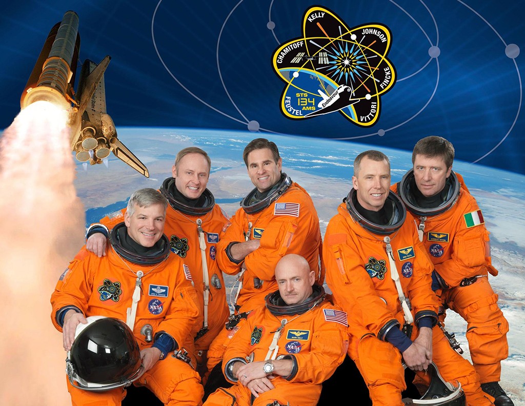 Seis astronautas sorridentes em trajes espaciais laranja posam com a Terra ao fundo, um &ocirc;nibus espacial decolando e o emblema da miss&atilde;o STS-134