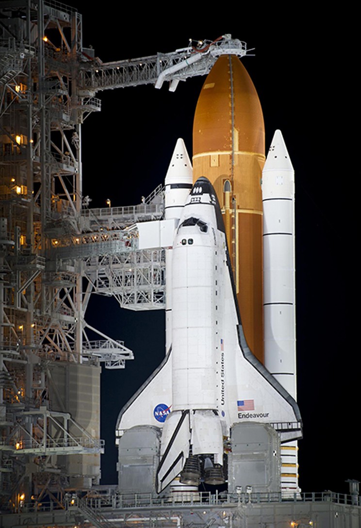 &Ocirc;nibus espacial Endeavour branco acoplado a um tanque de combust&iacute;vel laranja e dois propulsores brancos, em uma plataforma de lan&ccedil;amento iluminada &agrave; noite