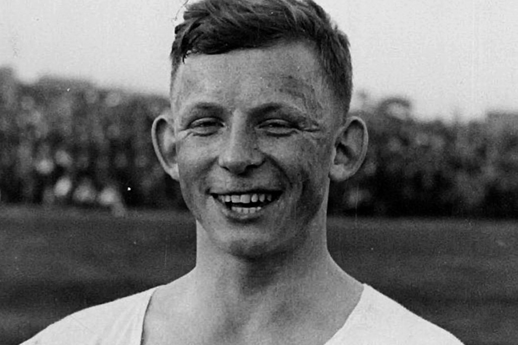 Homem jovem, branco, com cabelo curto e escuro, sorrindo amplamente com os dentes à mostra, vestindo uma camiseta clara, em um fundo desfocado de árvores e céu