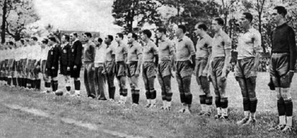 Time de futebol masculino em campo de grama, com jogadores e árbitros perfilados em duas fileiras, vestindo uniformes de época, em preto e branco