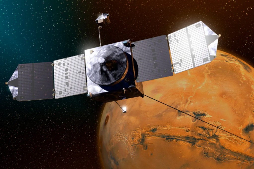 Sonda espacial cinza com painéis solares retangulares estendidos, orbitando o planeta Marte, que aparece em tons de laranja e marrom com crateras visíveis, em um fundo de espaço escuro com estrelas