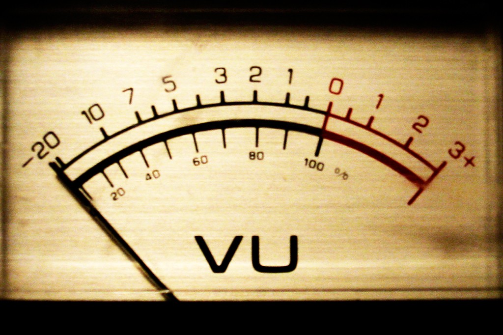 Medidor de VU analógico com ponteiro vermelho marcando a zona de sobrecarga, indicando volume alto. A escala superior vai de -20 a +3, e a inferior de 20% a 100%