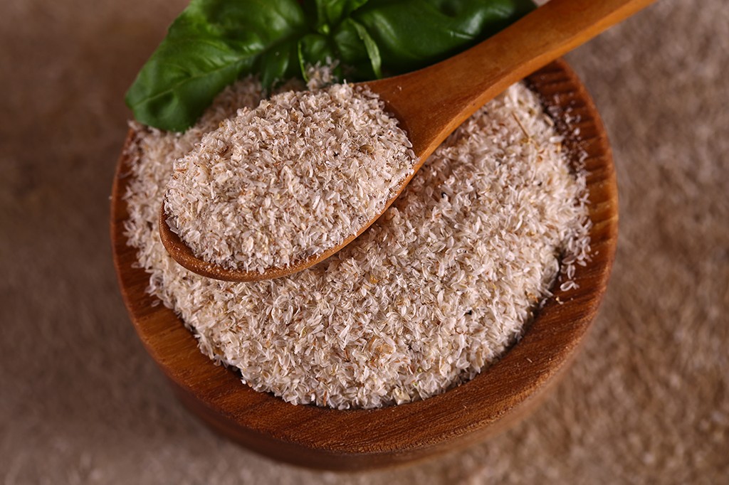 Fibras de psyllium em uma tigela de madeira com uma colher de pau cheia, e folhas verdes de manjericão ao fundo