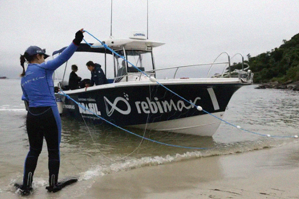 Mulher de roupa de mergulho azul e preta, com nadadeiras, puxa uma corda azul com boias brancas da &aacute;gua, em uma praia. Atr&aacute;s dela, uma lancha branca e preta com o logo 
