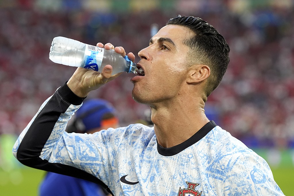 Cristiano Ronaldo, suado, bebe água de uma garrafa plástica transparente, vestindo camisa de manga comprida azul e branca com detalhes pretos, em um estádio lotado