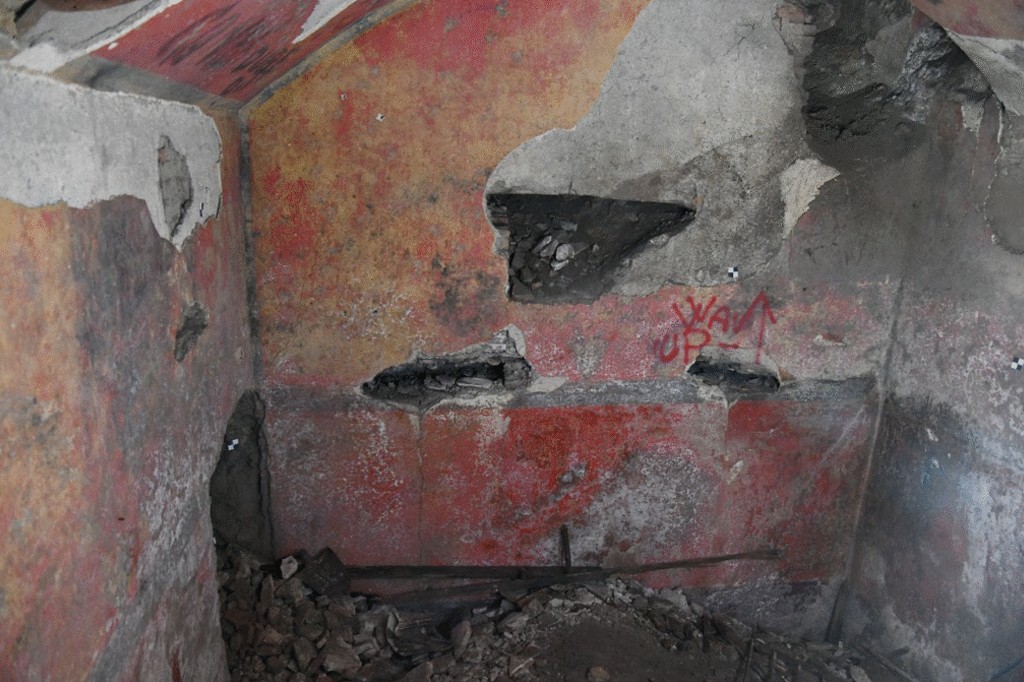 Interior de uma estrutura antiga com paredes vermelhas e amarelas descascadas, revelando concreto cinza e buracos. No ch&atilde;o, h&aacute; entulho de pedras e escombros. A parede central exibe a picha&ccedil;&atilde;o WAK UP em vermelho