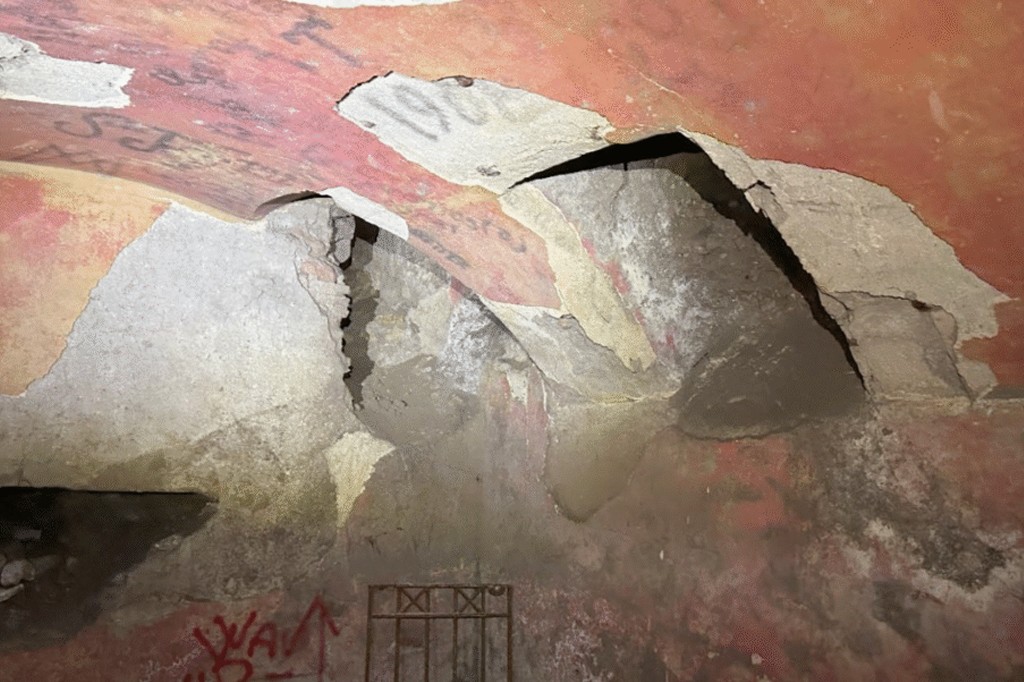 Teto abobadado de um t&uacute;nel subterr&acirc;neo com a pintura vermelha descascando e revelando concreto e tijolos. H&aacute; picha&ccedil;&otilde;es em preto e vermelho, e uma grade de metal enferrujada na parte inferior